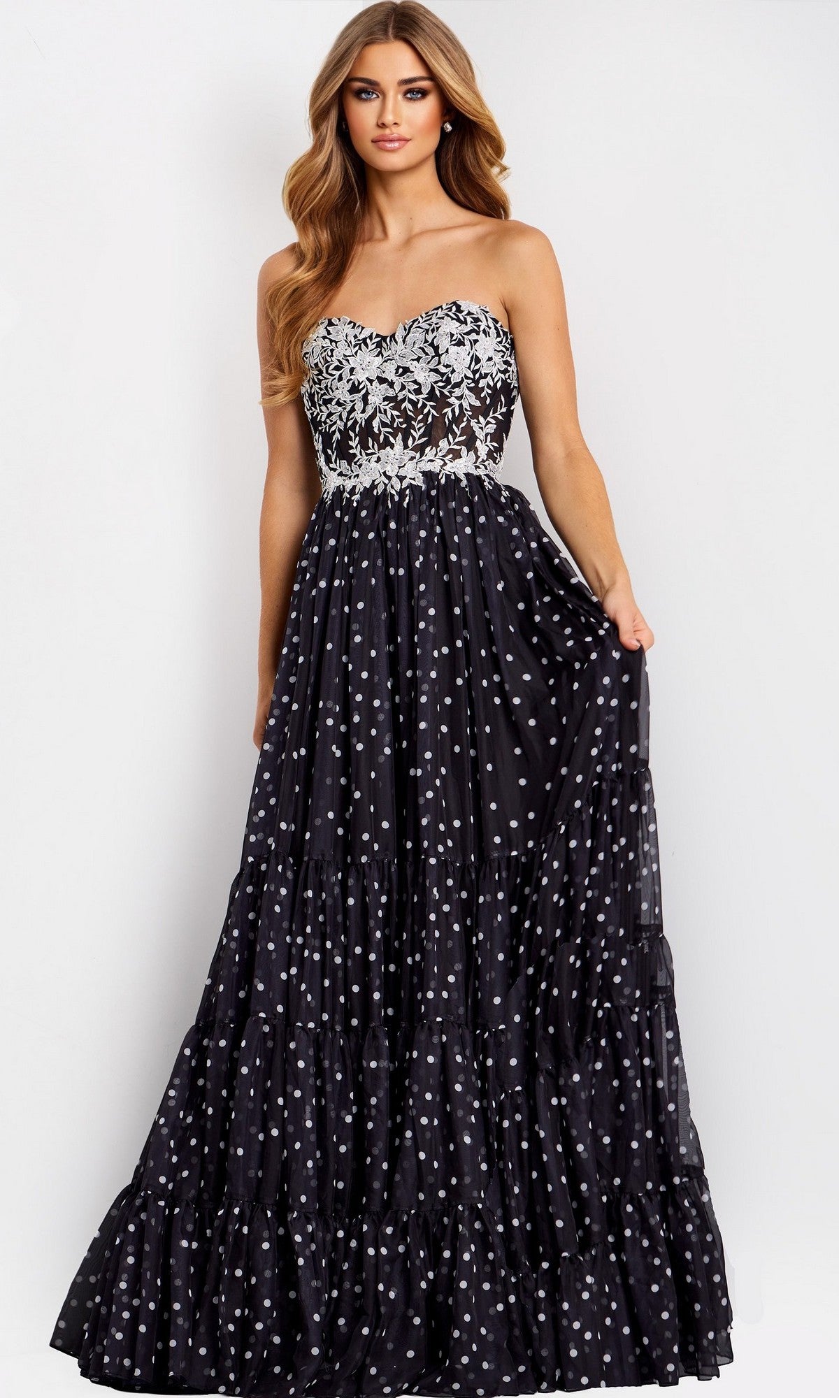 A woman models the Jovani 49086 long prom dress, showcasing a strapless sheer floral lace corset bodice and a full A-line polka dot skirt against a plain light background.